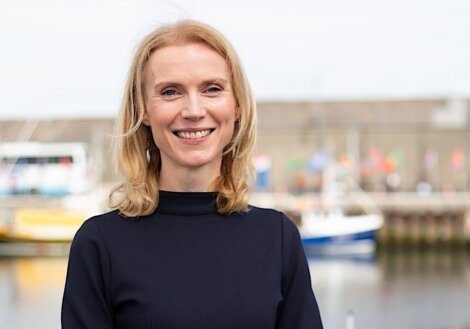 A woman with blonde hair and a black top stands smiling outdoors with boats and a waterfront visible in the background.