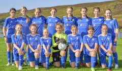 A girls' soccer team in blue uniforms poses on a grassy field with hills in the background. The goalkeeper, in green, holds a soccer ball.