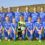 A girls' soccer team in blue uniforms poses on a grassy field with hills in the background. The goalkeeper, in green, holds a soccer ball.