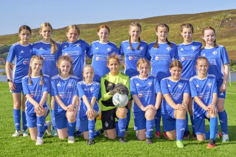 A girls' soccer team in blue uniforms poses on a grassy field with hills in the background. The goalkeeper, in green, holds a soccer ball.