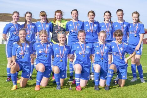 A women's soccer team in blue uniforms poses on a field with medals around their necks and a trophy in front of them.