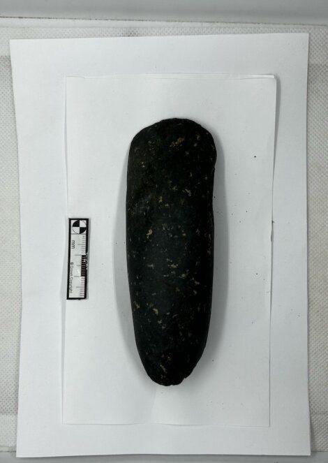 A black cylindrical stone artifact placed on a white paper with a measurement scale ruler for size reference.