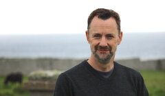 A man with short dark hair and a beard standing outdoors with a body of water and grassy area in the background. He is wearing a dark long-sleeve shirt.