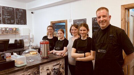 Five people are standing behind the counter of a café, wearing black aprons. There is a cake displayed in a glass dome and a hot water dispenser on the counter. Menu boards are on the wall behind them.