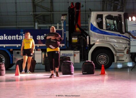 Two women in yellow shirts are carrying large bags in front of a parked truck indoors. The background includes safety cones and industrial equipment.