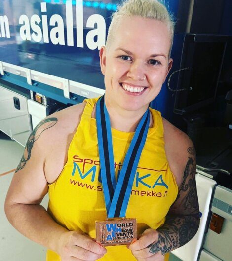 A person with short blonde hair, wearing a yellow tank top, holding a bronze medal on a blue ribbon, stands in front of a blue sign with partial text visible.