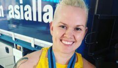 A person with short blonde hair and a nose ring smiles at the camera, wearing a yellow top and a medal around their neck. The background has a sign with partially visible text.