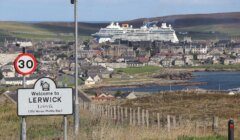 A road sign welcomes visitors to Lerwick, with a large cruise ship docked in the background and the town spread out along the coastline.