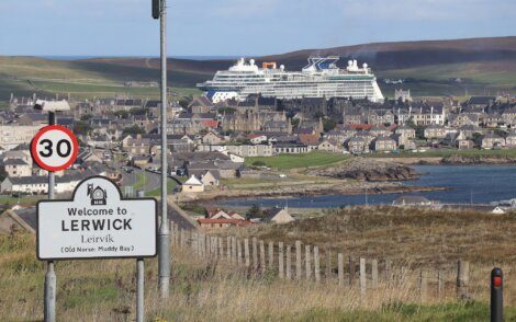 A road sign welcomes visitors to Lerwick, with a large cruise ship docked in the background and the town spread out along the coastline.