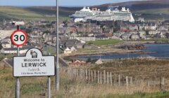 A road sign welcomes visitors to Lerwick, with a large cruise ship docked in the background and the town spread out along the coastline.