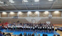 A large group of children perform a gymnastics routine in a gymnasium, with some standing and raising their arms. Spectators are seated in the foreground, watching the performance.