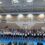 A large group of children perform a gymnastics routine in a gymnasium, with some standing and raising their arms. Spectators are seated in the foreground, watching the performance.