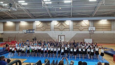 A large group of children perform a gymnastics routine in a gymnasium, with some standing and raising their arms. Spectators are seated in the foreground, watching the performance.