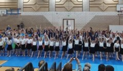 A large group of children perform a gymnastics routine in a gymnasium, with some standing and raising their arms. Spectators are seated in the foreground, watching the performance.