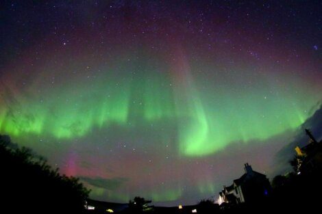 A night sky with green and pink aurora borealis above houses and trees, with stars visible in the background.