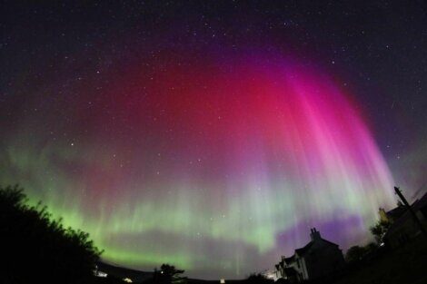 A night sky illuminated by a striking aurora display with vibrant shades of green and pink, visible above a silhouetted landscape with buildings and trees.