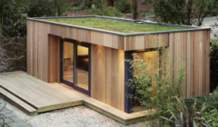 A modern wooden shed with large glass doors and a green roof, surrounded by a gravel area, wooden deck, and greenery.