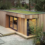 A modern wooden shed with large glass doors and a green roof, surrounded by a gravel area, wooden deck, and greenery.