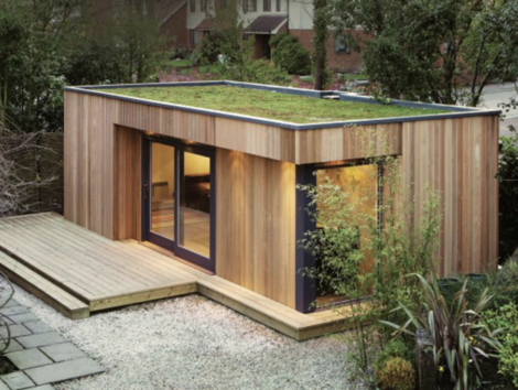 A modern wooden shed with large glass doors and a green roof, surrounded by a gravel area, wooden deck, and greenery.