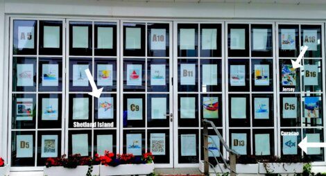 Window display featuring various artwork, each marked with a letter and number code. Labels "Shetland Island," "Jersey," and "Curacao" indicate origins of specific pieces. Red flowers line the bottom.