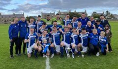 A soccer team in blue and white uniforms pose for a group photo on a field with trophies. Coaches and supporters stand alongside, while buildings and a cloudy sky are visible in the background.