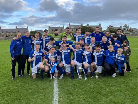 A soccer team in blue and white uniforms pose for a group photo on a field with trophies. Coaches and supporters stand alongside, while buildings and a cloudy sky are visible in the background.