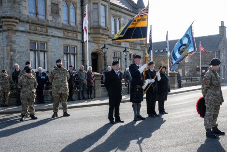 A group of people, some in military uniforms, stand in front of a large stone building with flags during a ceremony.