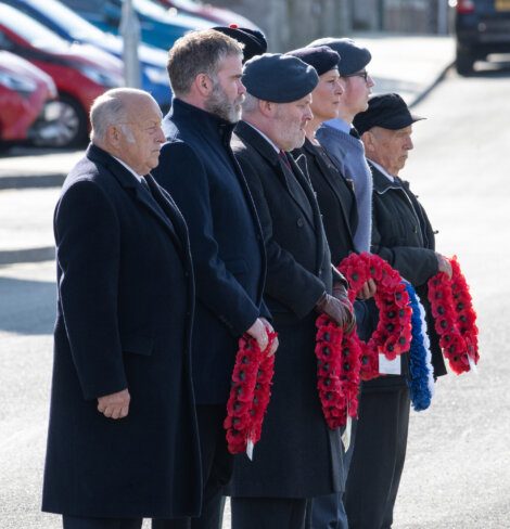 A group of six people stands in a line holding wreaths of red poppies during a remembrance event; five are wearing formal attire with berets or hats, and one person is in a suit without headgear.