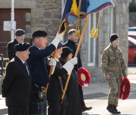 People holding flags and wreaths during a ceremony, with a person in military uniform in the background.