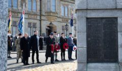 People stand in front of a memorial monument holding wreaths during a remembrance ceremony, with a building and military personnel in the background.