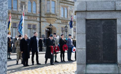 People stand in front of a memorial monument holding wreaths during a remembrance ceremony, with a building and military personnel in the background.