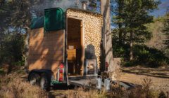 Mobile wooden sauna with a smoking chimney parked in a forest clearing.