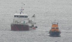 Two vessels at sea: a large boat with a white superstructure and a smaller orange lifeboat to its right.