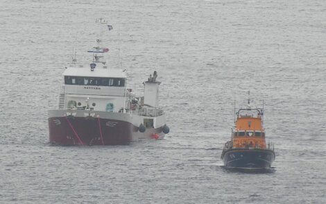 Two vessels at sea: a large boat with a white superstructure and a smaller orange lifeboat to its right.