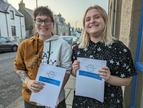 Two smiling individuals holding papers with the text "open project" on the sidewalk.