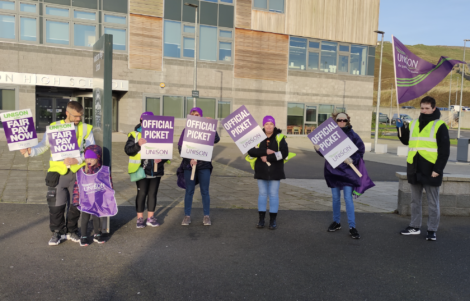 Group of individuals participating in a labor strike, holding signs for fair pay in front of a high school.