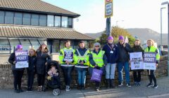 Group of people with signs on a picket line during a strike, wearing vests and displaying union affiliation.