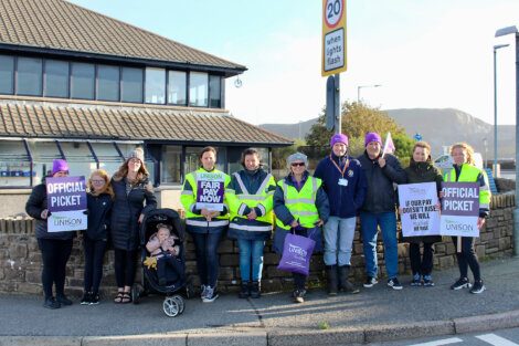 Group of people with signs on a picket line during a strike, wearing vests and displaying union affiliation.