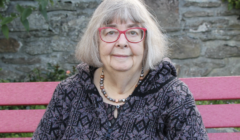 Woman with glasses sitting on a pink bench against a stone wall background.