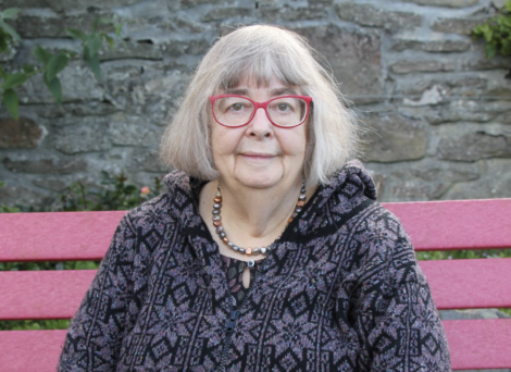 Woman with glasses sitting on a pink bench against a stone wall background.