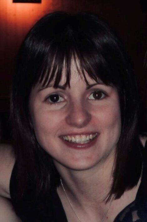 A woman with straight dark brown hair and bangs smiles at the camera. She is wearing a dark top and a necklace. The background is dimly lit with a warm light.