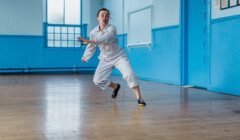 Person in white attire and black shoes performing a dynamic dance move in a spacious room with blue walls and windows.