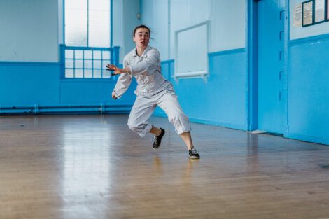 Person in white attire and black shoes performing a dynamic dance move in a spacious room with blue walls and windows.