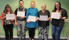 Five people standing in a row, each holding a certificate and smiling. They are in a room with a green wall and blue carpet.