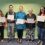 Five people standing in a row, each holding a certificate and smiling. They are in a room with a green wall and blue carpet.