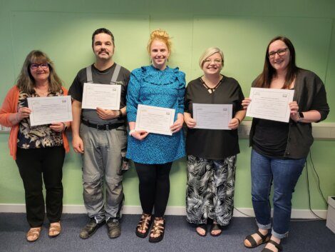 Five people standing in a row, each holding a certificate and smiling. They are in a room with a green wall and blue carpet.