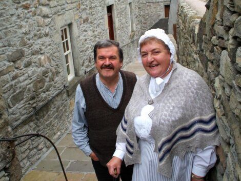 Two people in traditional clothing stand in a narrow stone alleyway. The woman wears a bonnet and shawl, while the man is in a vest and shirt. Stone walls and buildings are visible in the background.