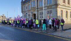 Group of individuals gathered for an event or demonstration on a city street, with some wearing high-visibility vests and carrying flags.