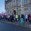 Group of individuals gathered for an event or demonstration on a city street, with some wearing high-visibility vests and carrying flags.