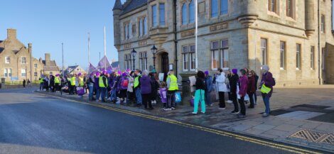 Group of individuals gathered for an event or demonstration on a city street, with some wearing high-visibility vests and carrying flags.